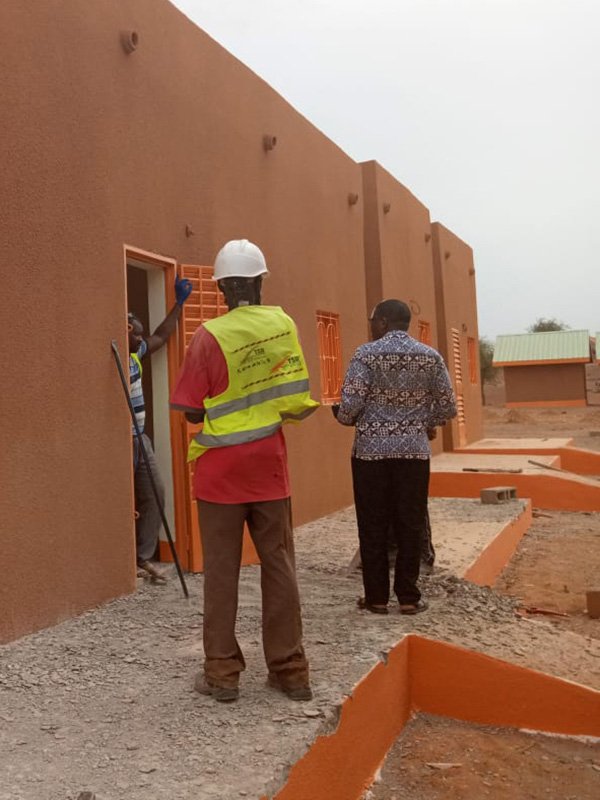 Construction site with individuals inspecting buildings, one wearing a safety vest and helmet, and another engaging with the structure.