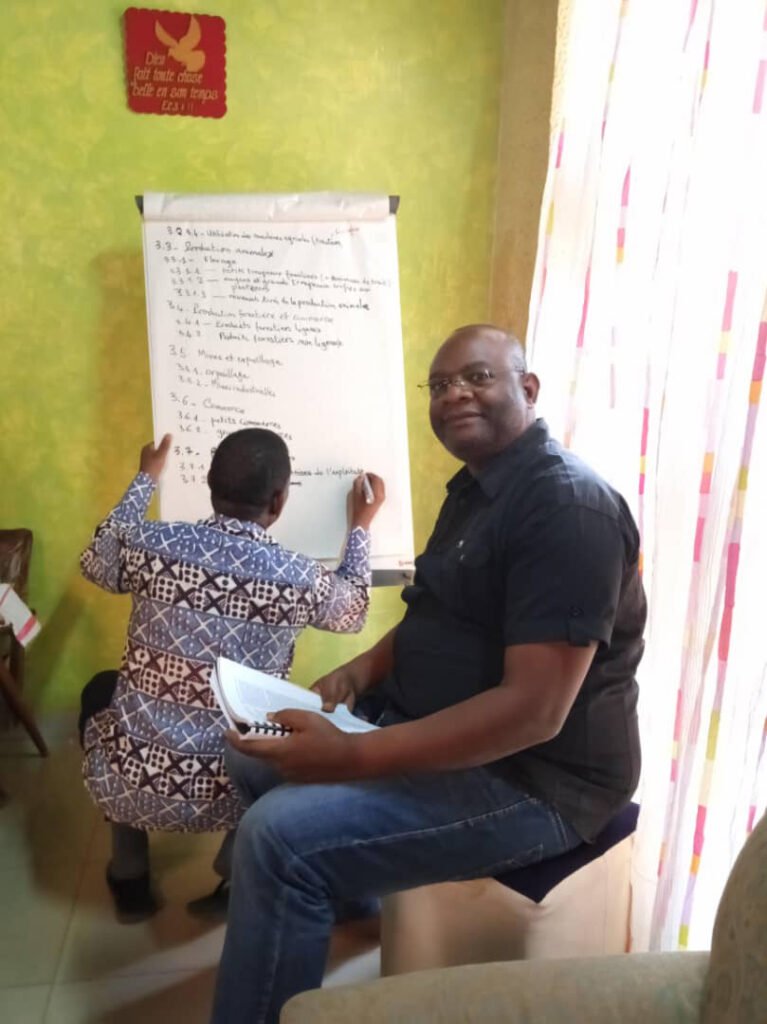 A man sitting with a notepad while another individual writes on a large paper board in a brightly lit room.
