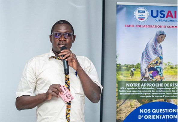A man speaking at an event, holding a card, with a banner displaying a woman in traditional attire and text about collaborative approaches.