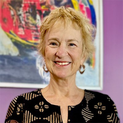A smiling woman with short blonde hair wearing a patterned black top, standing in front of a colorful abstract painting.