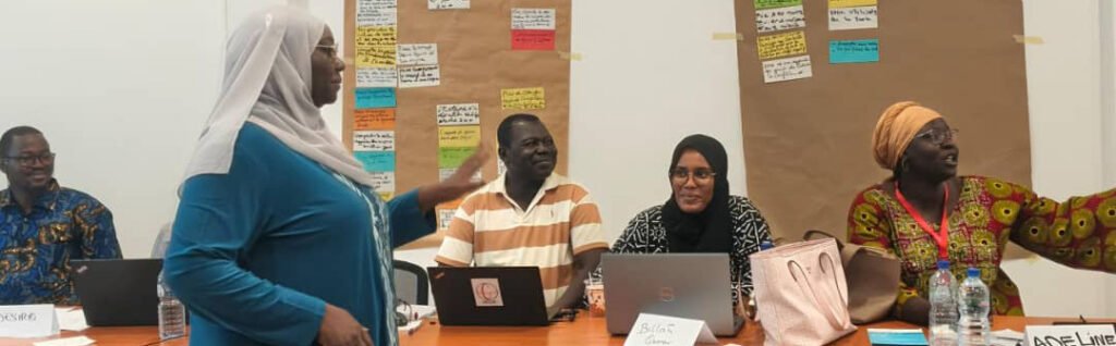 A group of people engaged in a discussion at a table, with laptops and colorful notes displayed on the wall behind them.