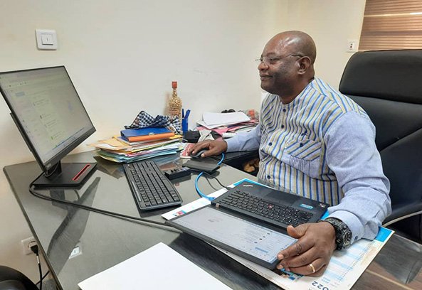 A man seated at a desk, working on a laptop and desktop computer, surrounded by paperwork and office supplies.
