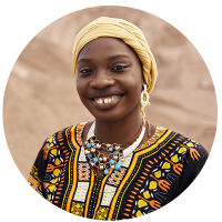 A smiling woman wears traditional attire with intricate patterns and jewelry against a sandy background.