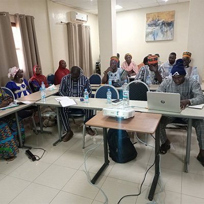A group of people seated at tables in a conference room, engaged in discussion, with a laptop and projector present.