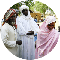 Three individuals in traditional attire, two men and a woman, engaged in conversation against a backdrop of simple structures and greenery.