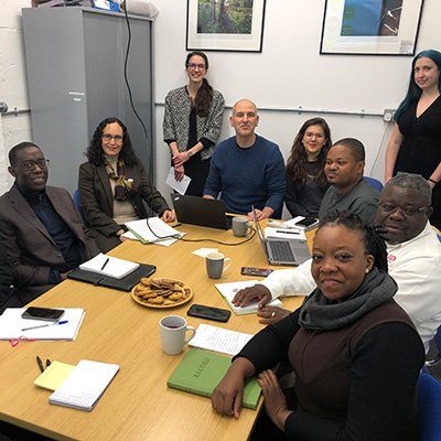 A diverse group of professionals gathered around a table, engaged in discussion with laptops and notebooks, sharing ideas and refreshments.
